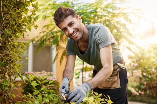 Close up outdoors portrait of beautiful cheerful bearded caucasian farmer in blue shirt and gloves smiling in camera, working with garden tools on his farm near countryside house